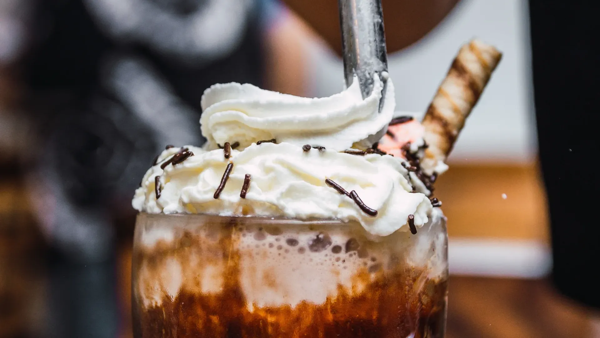Barista dispensing whipped cream from a stainless dispenser onto iced coffee drinks in a café