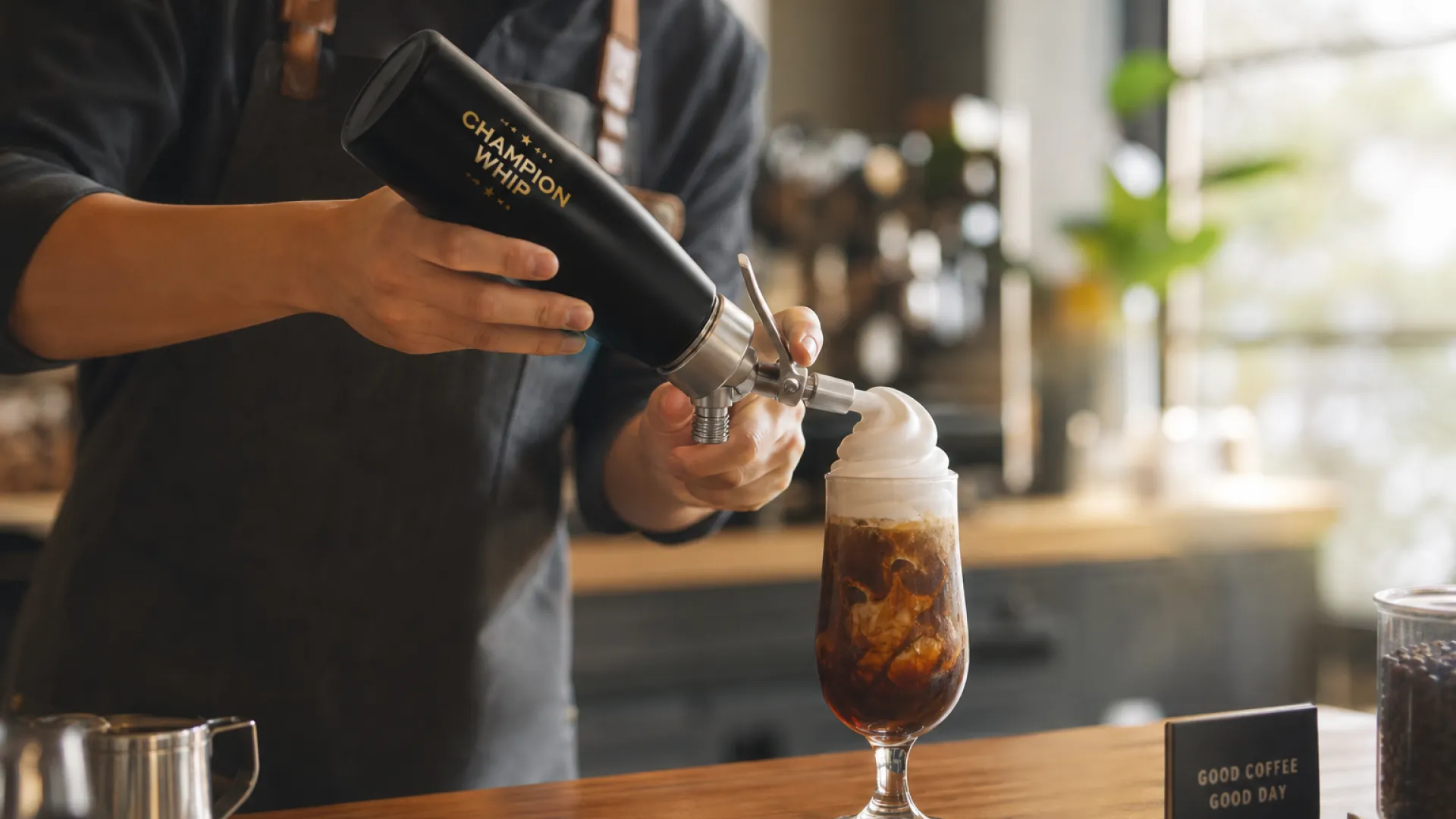 Barista topping an iced coffee with cold foam dispensed from a stainless steel cream whipper in a specialty coffee shop